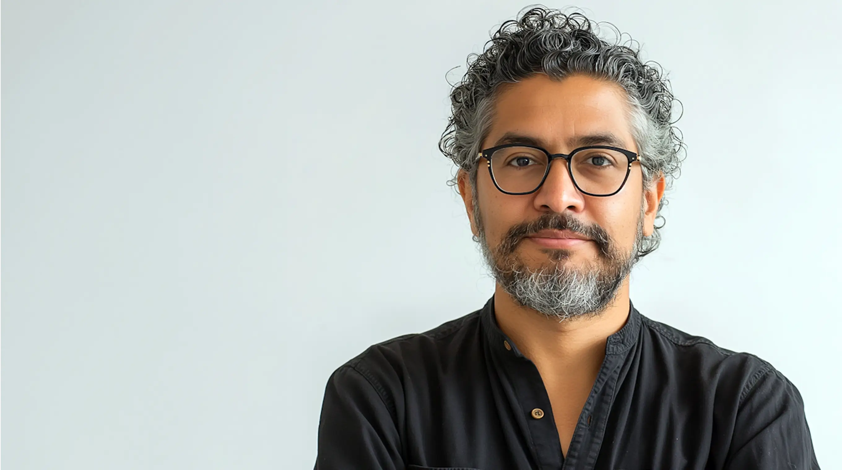 Man with curly salt-and-pepper hair and beard wearing black-rimmed glasses and a black shirt, smiling slightly against a plain light background.